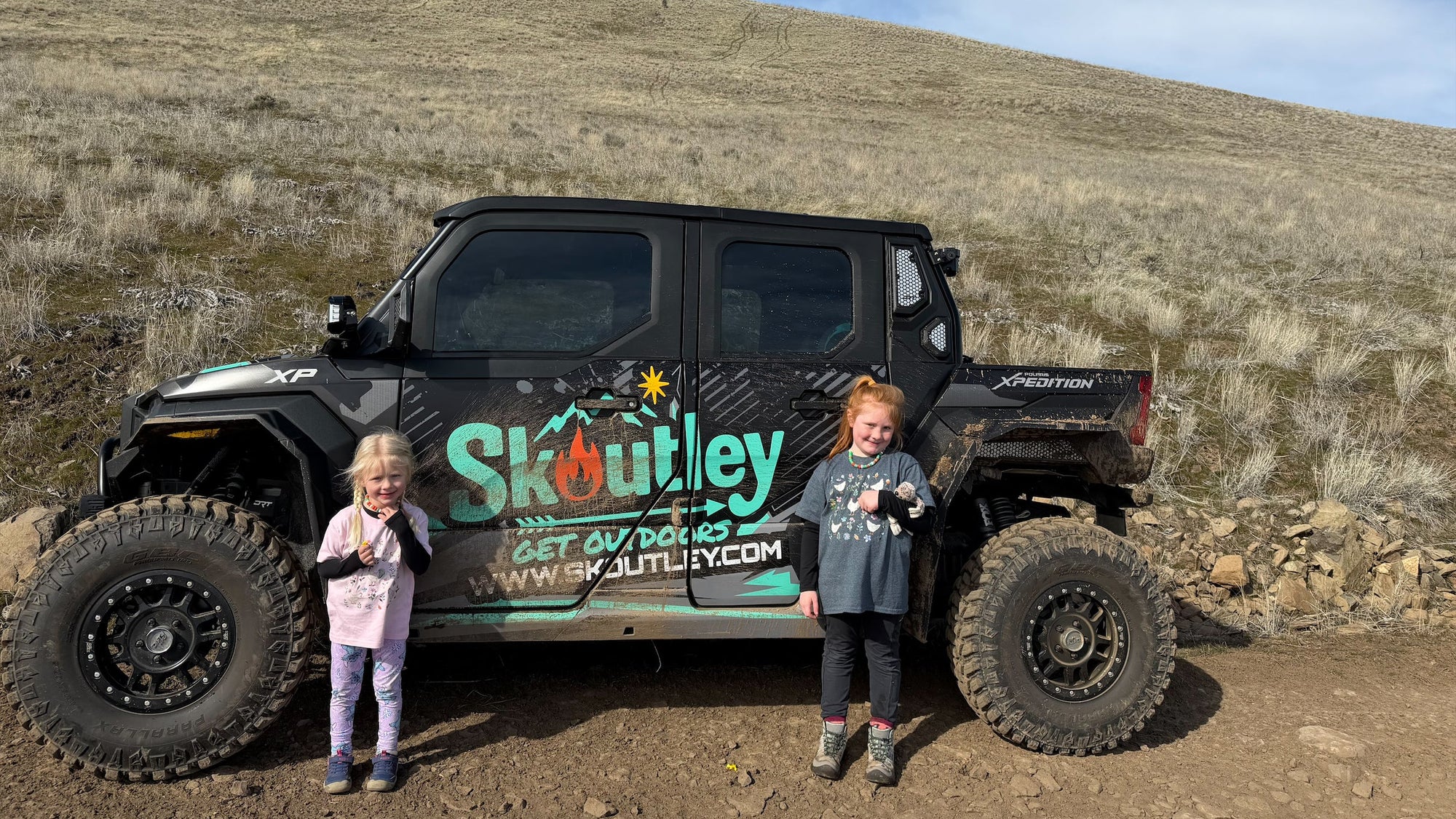 Two children standing next to a large off-road vehicle with 'Skoutley' branding on a rocky hillside.